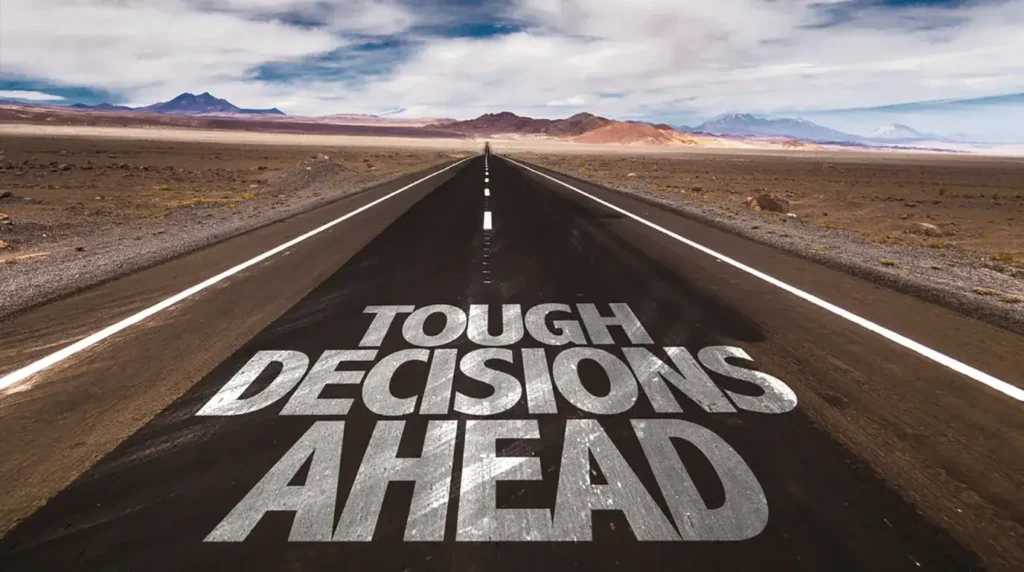 Teks "Tough Decisions Ahead" tertulis di jalan aspal kosong yang memanjang membelah lanskap padang pasir kering di bawah langit berawan.