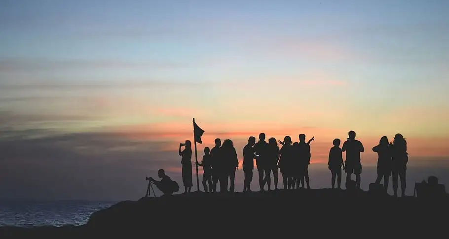 Siluet sekelompok orang berkumpul di tebing pantai saat matahari terbenam, ilustrasi dari file Orang-Orang-yang-Berebut.webp