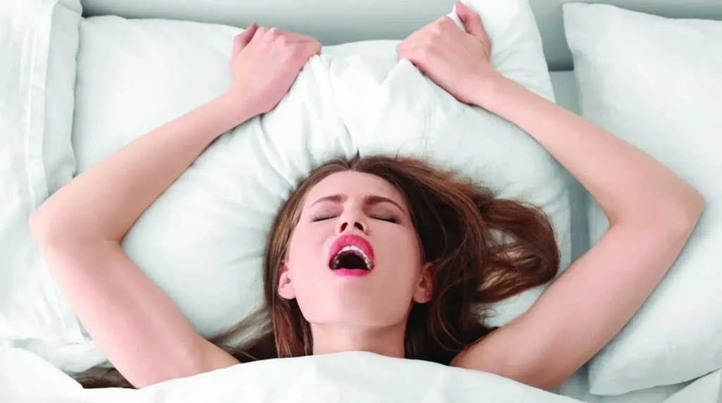 Ekspresi kepuasan seorang wanita saat tidur telentang di kasur putih sambil memegang bantal dengan kedua tangannya.