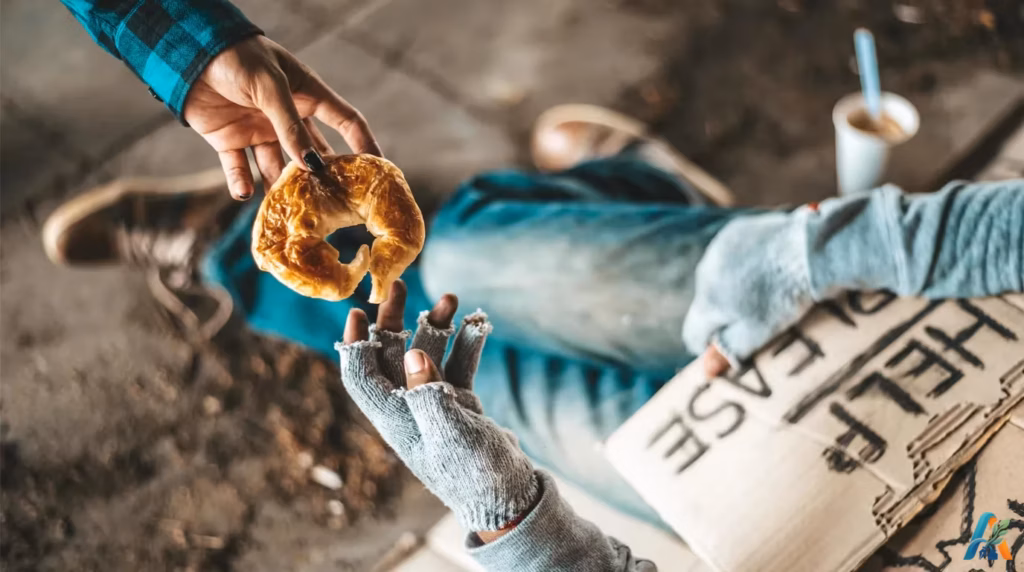 Tangan memberikan sebuah roti donat croissant kepada pengemis bersarung tangan rajut robek yang memegang kardus kosong.