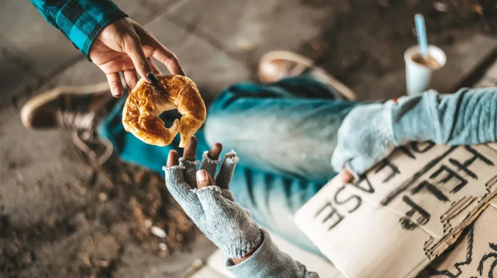 Tangan memberikan sebuah roti donat croissant kepada pengemis bersarung tangan rajut robek yang memegang kardus kosong.