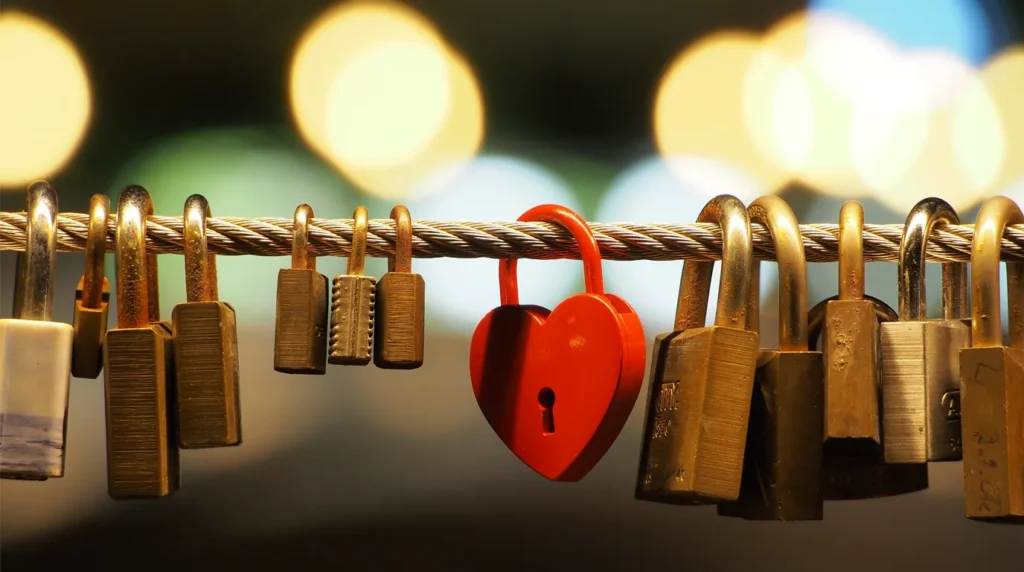 Gembok berbentuk hati warna merah di antara gembok-gembok kuningan yang dikaitkan pada kabel seling pagar.