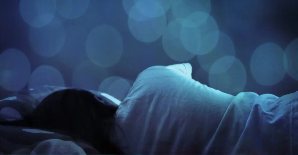 Seseorang tertidur pulas membelakangi kamera di bawah selimut dengan latar belakang bokeh biru, ilustrasi saat sepi dalam lelap