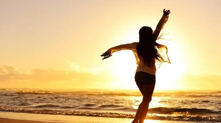 Siluet wanita berambut panjang merentangkan tangan di tepi pantai berlatar matahari terbenam yang jingga