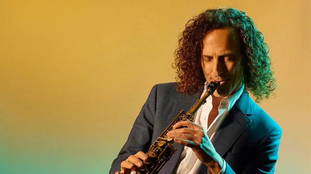 Pemain saksofon Kenny G berambut keriting panjang memegang saksofon dengan latar belakang kuning jingga.