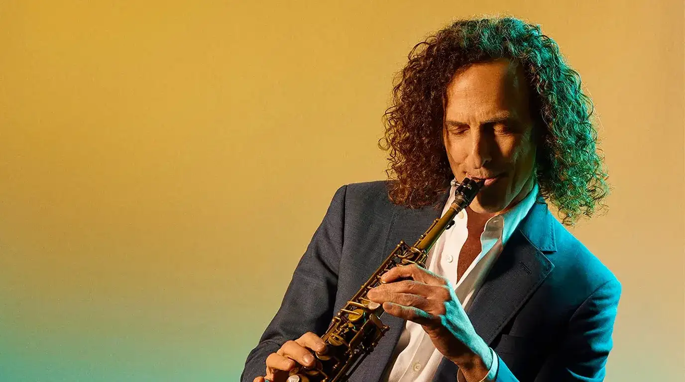 Pemain saksofon Kenny G berambut keriting panjang memegang saksofon dengan latar belakang kuning jingga.