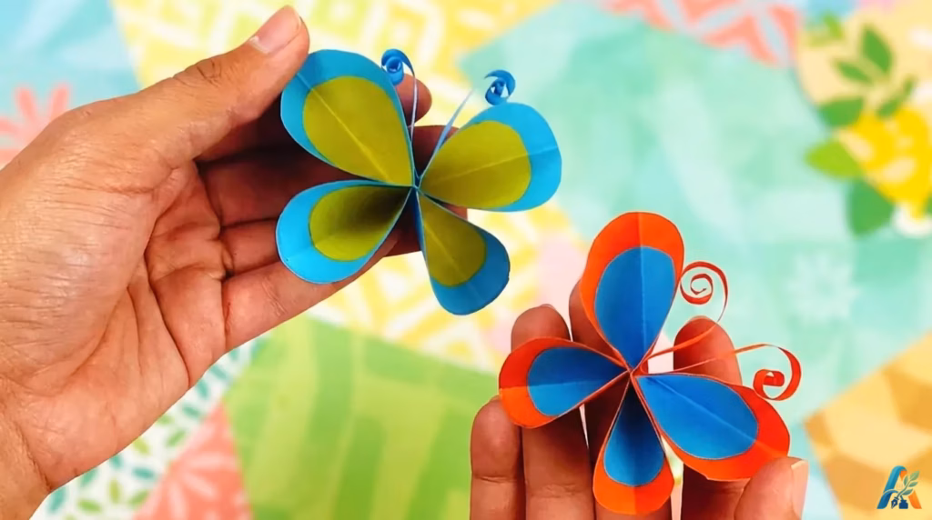 Dua kupu-kupu kertas origami biru, hijau, dan jingga dipegang tangan dengan latar belakang kertas warna-warni.