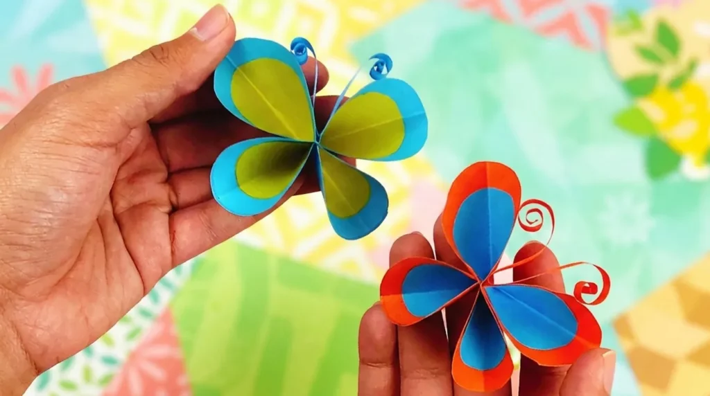 Dua kupu-kupu kertas origami biru, hijau, dan jingga dipegang tangan dengan latar belakang kertas warna-warni.