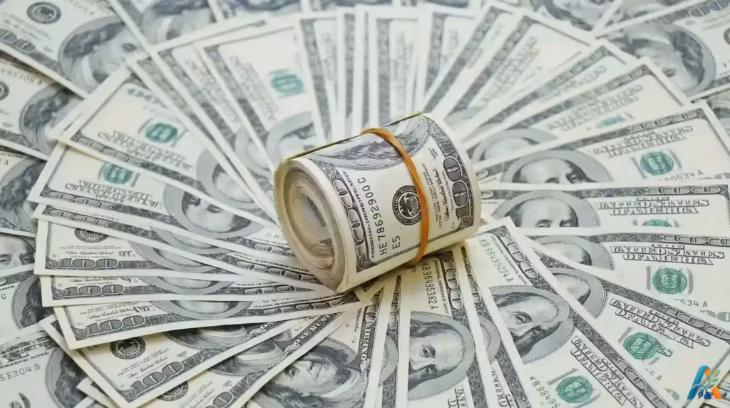 Gulungan uang kertas seratus dolar AS diletakkan di atas tumpukan uang kertas lainnya yang disusun membentuk kipas, melambangkan kekayaan finansial.