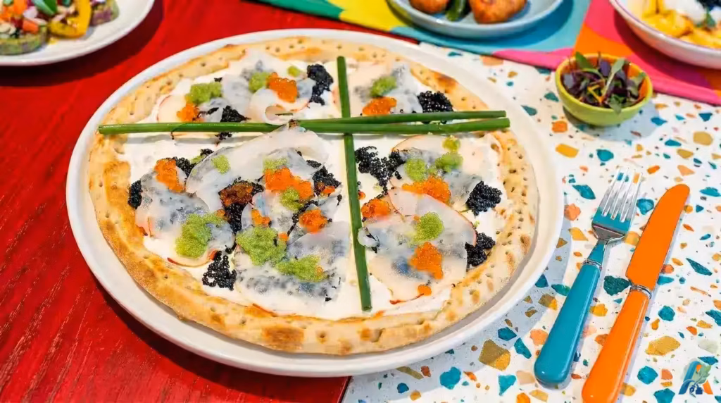 Nino's Bellissima Pizza beralaskan krim putih, kaviar hitam, telur ikan oranye, dan hiasan daun bawang di atas meja merah.