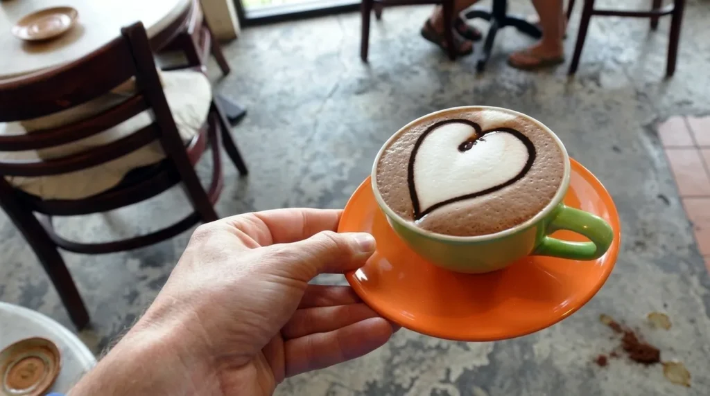 Seseorang memegang cangkir kopi berwarna hijau di atas tatakan oranye dengan seni latte berbentuk hati di dalam kafe.