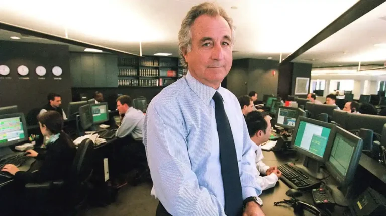 Bernard Madoff berdiri di depan meja kerjanya di lantai perdagangan yang sibuk, dikelilingi oleh karyawan dan layar komputer.