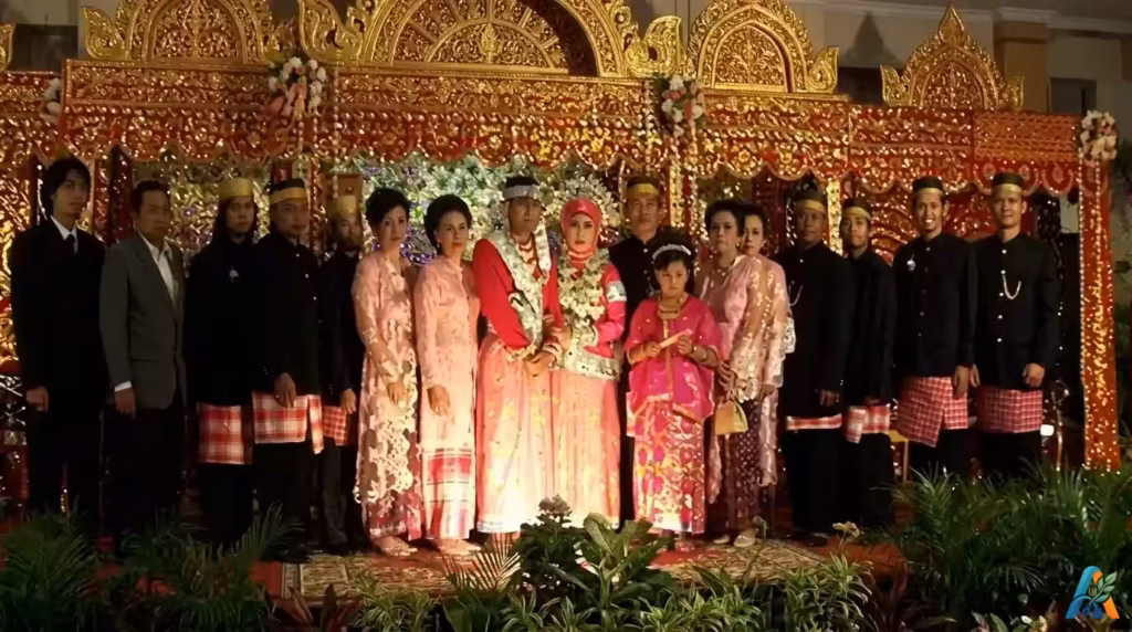 Pasangan pengantin Bugis mengenakan baju bodo tradisional berwarna merah muda di depan pelaminan megah penuh ukiran emas yang artistik.