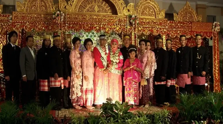 Pasangan pengantin Bugis mengenakan baju bodo tradisional berwarna merah muda di depan pelaminan megah penuh ukiran emas yang artistik.
