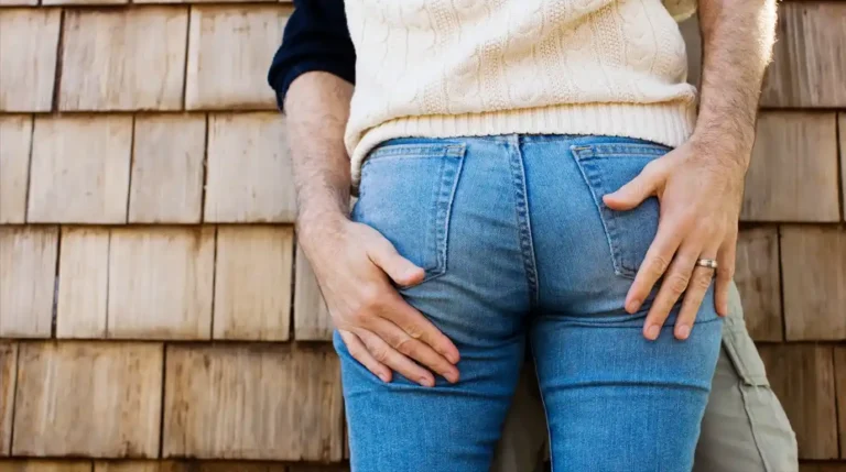 Pasangan mesra yang sedang berpelukan, terlihat dari jarak dekat fokus pada tangan yang menyentuh celana jeans, berlatar dinding kayu eksterior rumah.