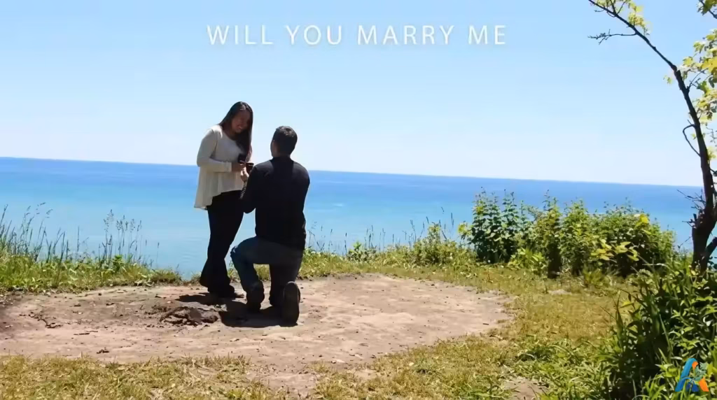 Seorang pria berlutut melamar kekasihnya di tebing tepi laut yang indah dengan teks "WILL YOU MARRY ME" di atasnya, dari file will-you-marry-me.webp.