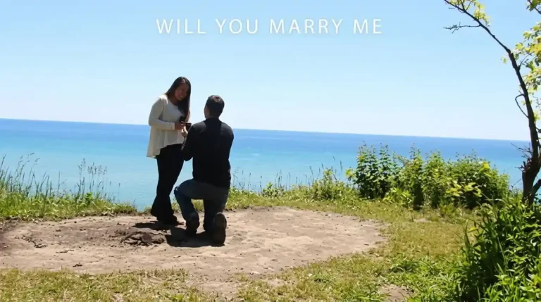 Seorang pria berlutut melamar kekasihnya di tebing tepi laut yang indah dengan teks "WILL YOU MARRY ME" di atasnya, dari file will-you-marry-me.webp.