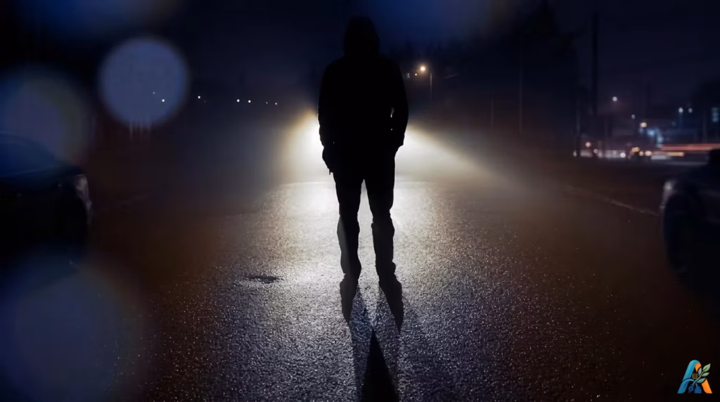 Siluet seorang pria berdiri di tengah jalan saat malam hari, diterangi oleh sorot lampu kendaraan dari belakang, sebagai ilustrasi puitis untuk tema dunia-ini-bagiku-hanyalah-tempatku-menunggu-mati.webp.