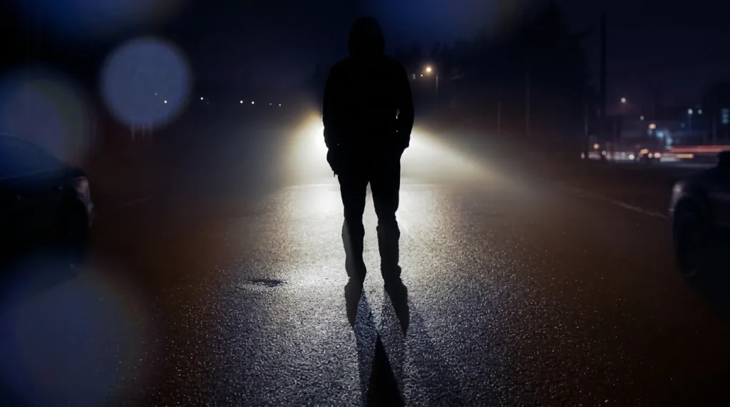 Siluet seorang pria berdiri di tengah jalan saat malam hari, diterangi oleh sorot lampu kendaraan dari belakang, sebagai ilustrasi puitis untuk tema dunia-ini-bagiku-hanyalah-tempatku-menunggu-mati.webp.