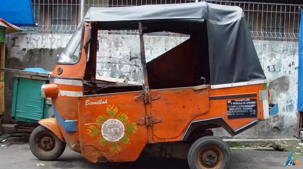 Bajaj oranye khas Jakarta tampak samping terparkir di pinggir jalan dekat dinding semen usang