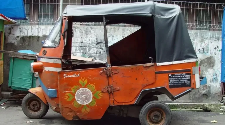 Bajaj oranye khas Jakarta tampak samping terparkir di pinggir jalan dekat dinding semen usang