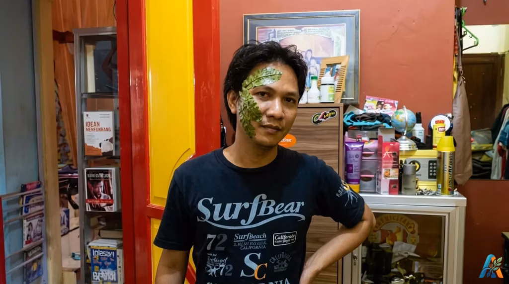 Seorang pria dengan daun hijau di wajahnya sebagai pengobatan tradisional untuk sakit gigi, di dalam sebuah rumah.