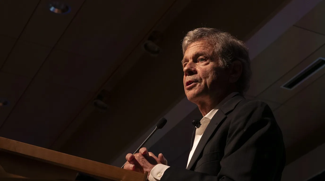 Alan Lightman memberikan ceramah atau kuliah umum di balik podium dengan jas hitam