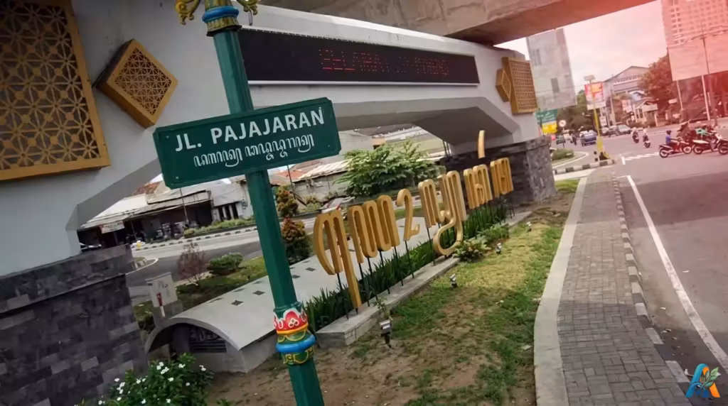 Plang nama jalan Jl. Pajajaran dengan tulisan aksara Jawa di Yogyakarta, menampilkan suasana kota dengan nuansa budaya lokal yang khas.