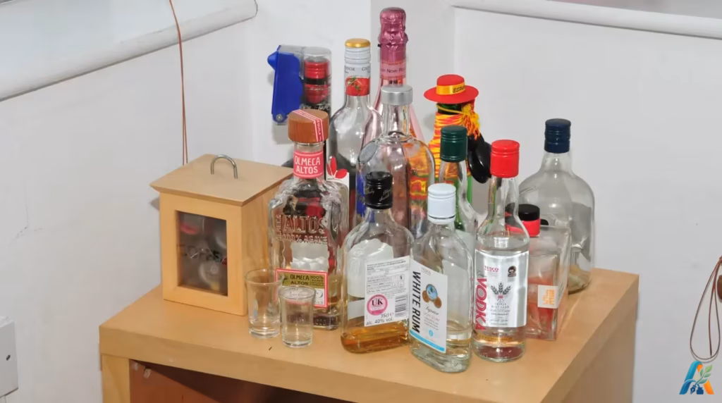 Koleksi botol minuman beralkohol di apartemen Sinaga, termasuk vodka, rum, tequila, dan anggur, tertata di atas meja samping kayu.