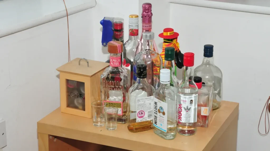 Koleksi botol minuman beralkohol di apartemen Sinaga, termasuk vodka, rum, tequila, dan anggur, tertata di atas meja samping kayu.
