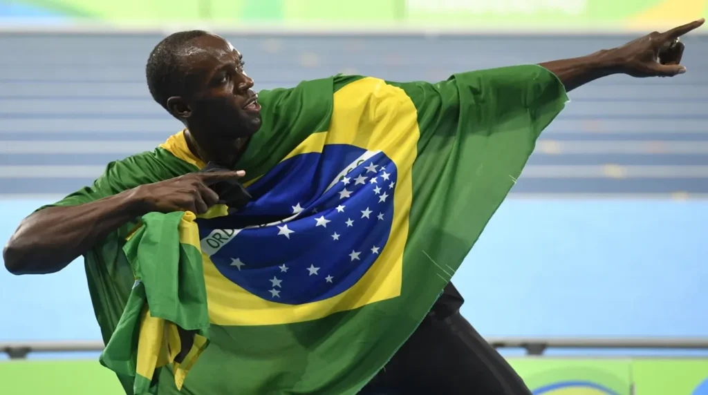 Usain Bolt merayakan kemenangan di lintasan lari dengan menyampirkan bendera Brasil di bahunya dan berpose ikonik menunjuk ke atas.
