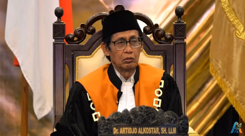 Dr. Artidjo Alkostar, S.H., LL.M., duduk di kursi hakim mengenakan toga kebesaran dengan latar belakang bendera dan dekorasi formal