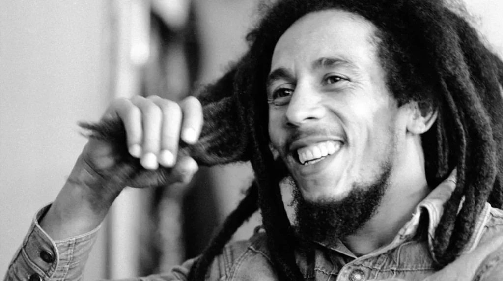 Potret hitam putih ikonik Bob Marley tersenyum sambil memegang rambut gimbalnya, menginspirasi kekuatan dan ketangguhan hidup