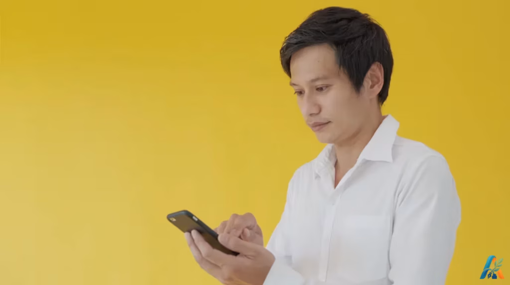 Seorang pria Asia mengenakan kemeja putih sedang serius menggunakan smartphone dengan latar belakang kuning polos