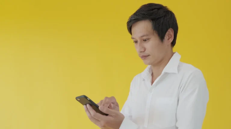 Seorang pria Asia mengenakan kemeja putih sedang serius menggunakan smartphone dengan latar belakang kuning polos
