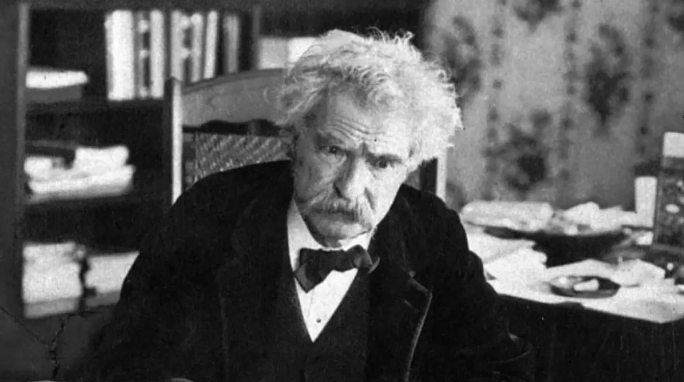 Potret hitam putih Mark Twain duduk di ruang kerja dengan rambut putih ikal khasnya