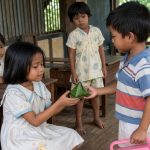 Ilustrasi anak-anak TK di Merauke tahun 1980-an, seorang anak laki-laki memberikan koci-koci berbungkus daun pisang kepada seorang anak perempuan pendiam di ruang kelas sederhana TK Al Hikmah Spadem.