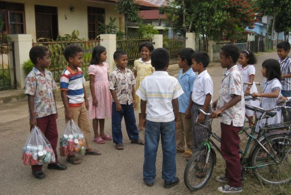 Sekelompok anak-anak Papua dan non-Papua berdiri rame-rame di jalan perumahan Merauke pada hari Lebaran, memakai baju baru, sebagian pegang kantong plastik berisi minuman kaleng, dengan sepeda dan rumah-rumah modern tahun 1980-an di latar belakang. Kalau ko mau versi yang sedikit lebih pendek untuk blog: Anak-anak di Merauke saat Lebaran berdiri rame-rame di jalan perumahan, memakai baju baru dan membawa kantong plastik berisi minuman kaleng.